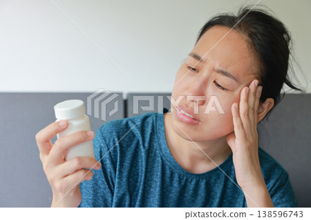 Sick Asian woman suffering from severe headache or migraine touching her head in pain while looking at a white pill bottle. Healthcare, sickness, stress, medical treatment and painkiller concept. Sick Asian woman suffering from severe headache or migraine touching her head in pain while looking at a white pill bottle. Healthcare, sickness, stress, medical treatment and painkiller concept. 138596743
