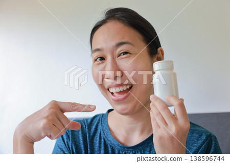 Happy smiling Asian woman pointing finger at a blank white pill bottle. Positive healthcare, medicine, vitamin, dietary supplement recommendation, mockup advertising and illness recovery concept. Happy smiling Asian woman pointing finger at a blank white pill bottle. Positive healthcare, medicine, vitamin, dietary supplement recommendation, mockup advertising and illness recovery concept. 138596744