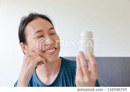 Happy smiling Asian woman looking fondly at a blank white pill bottle. Positive healthcare, collagen, vitamin, beauty supplement or medicine product mockup advertising and treatment recovery concept. Happy smiling Asian woman looking fondly at a blank white pill bottle. Positive healthcare, collagen, vitamin, beauty supplement or medicine product mockup advertising and treatment recovery concept. 138596745