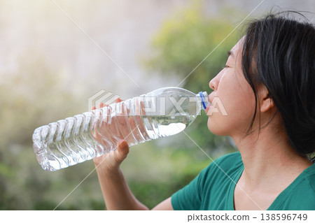 Close up side view of young Asian woman drinking pure mineral water from a clear plastic bottle outdoors. Healthy hydration, quenching thirst, wellness, body refreshment and summer lifestyle concept. 138596749