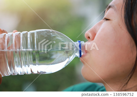 Close up side view of young Asian woman drinking pure mineral water from a clear plastic bottle outdoors. Healthy hydration, quenching thirst, wellness, body refreshment and summer lifestyle concept. 138596752