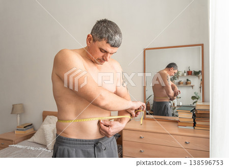 Man measuring his waist with a tape measure, checking progress on his weight loss journey in the bedroom. Real person, self-acceptance, authentic lifestyle Man measuring his waist with a tape measure, checking progress on his weight loss journey in the bedroom. Real person, self-acceptance, authentic lifestyle 138596753