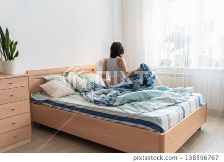 Woman sitting up in bed in a bright bedroom, starting the morning and looking towards the daylight from the window. The ordinary life of a real person. Authentic lifestyle Woman sitting up in bed in a bright bedroom, starting the morning and looking towards the daylight from the window. The ordinary life of a real person. Authentic lifestyle 138596767