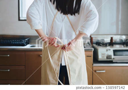 A woman putting on an apron in the kitchen; a scene from everyday life involving housework and cooking. A woman putting on an apron in the kitchen; a scene from everyday life involving housework and cooking. 138597142