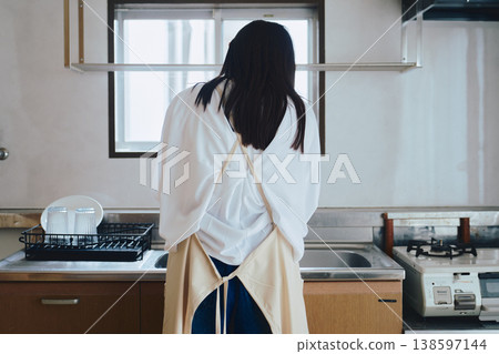 A woman putting on an apron in the kitchen; a scene from everyday life involving housework and cooking. A woman putting on an apron in the kitchen; a scene from everyday life involving housework and cooking. 138597144