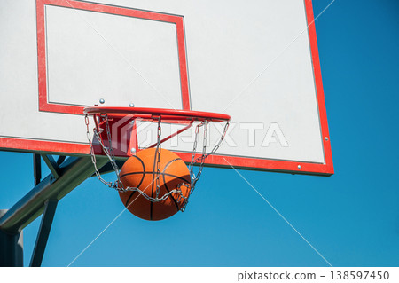 Basketball ball scoring through hoop with chain net against blue sky 138597450