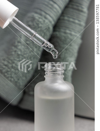 Serum drop falling from pipette into glass bottle against sage green bath towel. Cosmetic treatment 138597781