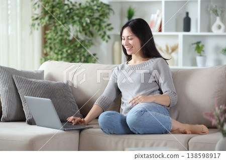 Happy asian woman is using laptop sitting on a couch Happy asian woman is using laptop sitting on a couch 138598917
