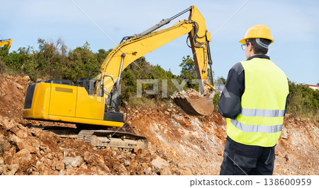 Engineer in a helmet with a digital tablet stands next to construction excavator 138600959