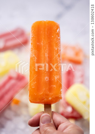 Hand holding a orange popsicle on ice pops and colorful popsicles background Hand holding a orange popsicle on ice pops and colorful popsicles background 138602418