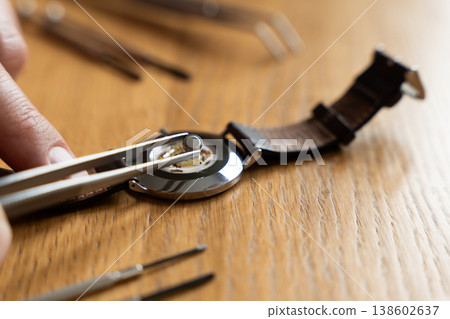 Close-up of a hand using tweezers to replace a watch battery on a wooden surface with watch tools nearby Close-up of a hand using tweezers to replace a watch battery on a wooden surface with watch tools nearby 138602637