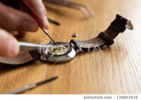 Close-up of a person using tweezers to repair a watch movement on a wooden workbench, with tools visible in the background Close-up of a person using tweezers to repair a watch movement on a wooden workbench, with tools visible in the background 138602638