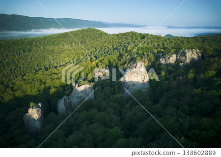 Aerial view of rocky formation surrounded by dense, verdant forest. Morning mist lingers over distant hills, creating serene and mystical atmosphere. Dovbush Rocks, Carpathian mountains, Ukraine. Aerial view of rocky formation surrounded by dense, verdant forest. Morning mist lingers over distant hills, creating serene and mystical atmosphere. Dovbush Rocks, Carpathian mountains, Ukraine. 138602889