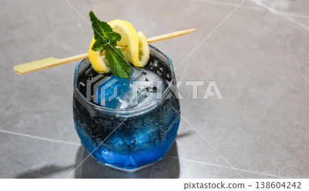close up on a blue cocktail with large rock ice, lemon slice and lime on top in a glass on a grey marble table with copy space close up on a blue cocktail with large rock ice, lemon slice and lime on top in a glass on a grey marble table with copy space 138604242