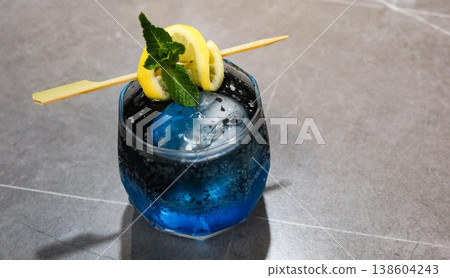 close up on a blue cocktail with large rock ice, lemon slice and lime on top in a glass on a grey marble table with copy space 138604243