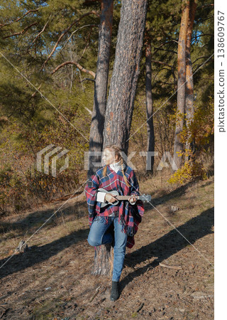 Portrait of smiling caucasian girl plays ukulele in autumnal park. Young woman plays guitar musical instrument outside in nature fall time. Audio music healing 138609767