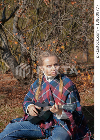Beautiful blonde girl playing guitar ukulele and singing relaxing enjoying outdoors on sunny autumn day. Hippie woman in boho style training playing musical instrument. Fall leaves 138609777