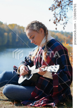 Portrait of smiling caucasian girl plays ukulele in autumnal park. Young woman plays guitar musical instrument outside in nature fall time. Audio music healing Portrait of smiling caucasian girl plays ukulele in autumnal park. Young woman plays guitar musical instrument outside in nature fall time. Audio music healing 138609781