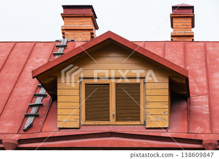 Yellow wooden dormer with closed louvered window set on a red metal roof 138609847