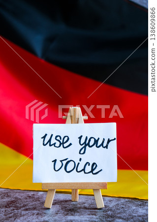 Paper note with words USE YOUR VOICE on National symbol of Germany. German wave silky flag background. Ellection political participation campaign and civic engagement Paper note with words USE YOUR VOICE on National symbol of Germany. German wave silky flag background. Ellection political participation campaign and civic engagement 138609866