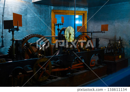 Intricate clockwork mechanism with gears and clock face in a dimly lit room with blue walls and wooden window frame 138611134