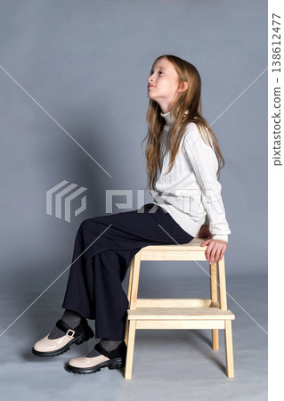 Girl sits wooden step stool with hands gently placed on her knee, subtle smile and quiet confidence Girl sits wooden step stool with hands gently placed on her knee, subtle smile and quiet confidence 138612477
