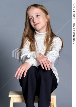 Girl sits wooden step stool with hands gently placed on her knee, subtle smile and quiet confidence 138612478
