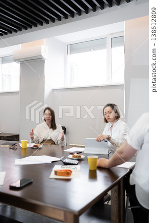 Colleagues conversing and eating at light wooden table after breakfast meeting in bright office. Conversing, eating, breakfast, wooden table, office. 138612809