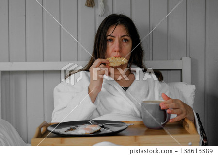 A calm woman enjoys a quiet breakfast. A serene lady sits in bed in a robe, holding a mug and a cracker, in the cozy atmosphere of her home. Morning rituals alone. A cozy interior. Copy space. Banner 138613389
