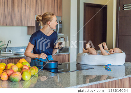 Mother talking to baby lying in cozy lounger on kitchen table while cooking at home. Caring mom interacting with infant during daily routine. Parenting, multitasking and family life concept Mother talking to baby lying in cozy lounger on kitchen table while cooking at home. Caring mom interacting with infant during daily routine. Parenting, multitasking and family life concept 138614064