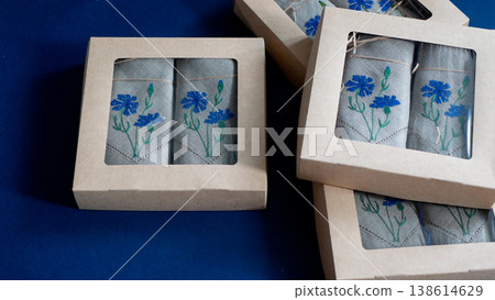 Linen napkins featuring delicate blue cornflower embroidery in eco-friendly craft paper boxes. Elegant embroidered textile accessories Linen napkins featuring delicate blue cornflower embroidery in eco-friendly craft paper boxes. Elegant embroidered textile accessories 138614629