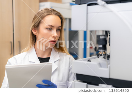 Performing data evaluation while holding documentation sheets at high tech laboratory workstation. Professional lab environment with advanced analytical instruments and clean surfaces visible. 138614890