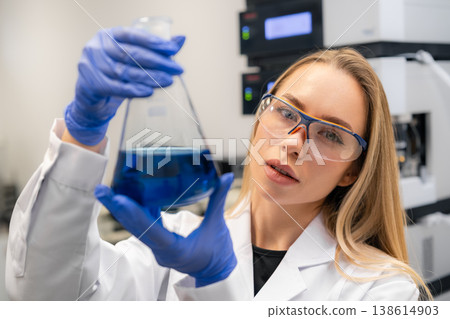 A researcher evaluates blue chemical solution within a laboratory flask at an advanced analytical testing facility. A researcher evaluates blue chemical solution within a laboratory flask at an advanced analytical testing facility. 138614903