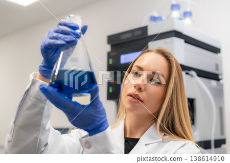 Analytical chemist inspecting a blue liquid in a laboratory flask while wearing gloves. The clean lab environment features advanced analytical instruments and equipment in the background. Analytical chemist inspecting a blue liquid in a laboratory flask while wearing gloves. The clean lab environment features advanced analytical instruments and equipment in the background. 138614910