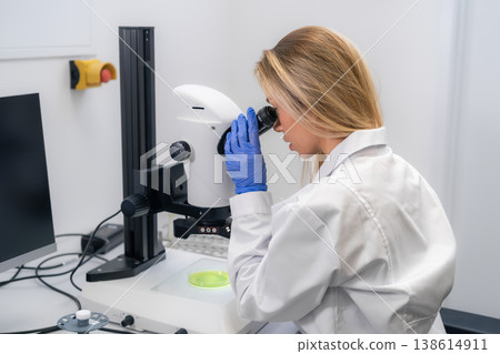 Laboratory technician examining green biological specimens under high powered microscope at research facility. Clean workspace with digital displays and precise instruments visible in background. Laboratory technician examining green biological specimens under high powered microscope at research facility. Clean workspace with digital displays and precise instruments visible in background. 138614911