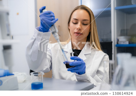 Technician using a micropipette to transfer liquid from one container to another at laboratory workstation. The workspace features various glassware and a clean, organized environment. 138614924