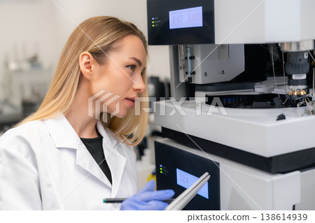 Technician monitoring advanced analytical system HPLC during biological sample analysis in clinical lab environment. Well lit workspace with various laboratory equipment visible in the background. 138614939