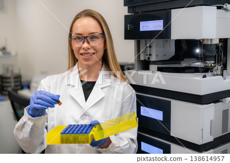 Clinical technician operating precision automated HPLC analyzer while holding amber vial and yellow microcentrifuge tube rack at laboratory workstation. Clinical technician operating precision automated HPLC analyzer while holding amber vial and yellow microcentrifuge tube rack at laboratory workstation. 138614957