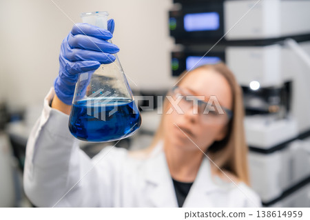 Laboratory technician inspecting blue colored liquid in Erlenmeyer flask while wearing protective gloves and eyewear. 138614959