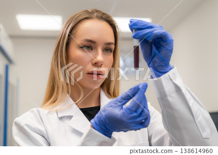 Laboratory technician analyzing a blood sample contained within a test tube at a clinical research facility. Clean, well lit environment with sterile surfaces and laboratory equipment visible. Laboratory technician analyzing a blood sample contained within a test tube at a clinical research facility. Clean, well lit environment with sterile surfaces and laboratory equipment visible. 138614960