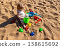 Toddler sitting on sandy beach playing with plastic bucket shovel and sand molds. Outdoor beach photography. Childhood and summer vacation concept. Design for poster, banner, postcard. Top view shot 138615692