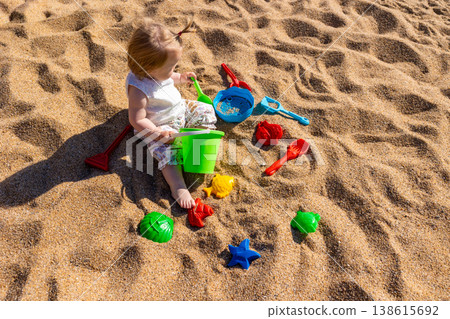 Toddler sitting on sandy beach playing with plastic bucket shovel and sand molds. Outdoor beach photography. Childhood and summer vacation concept. Design for poster, banner, postcard. Top view shot 138615692