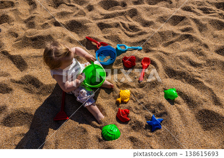 Toddler sitting on sandy beach playing with plastic bucket shovel and sand molds. Outdoor beach photography. Childhood and summer vacation concept. Design for poster, banner, postcard. Top view shot 138615693
