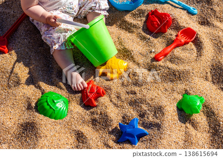 Toddler playing with plastic bucket shovel and sand molds on sandy beach. Outdoor beach photography. Childhood leisure and summer vacation concept. Design for poster, banner, postcard. Top view shot 138615694