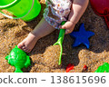 Toddler hand holding green shovel with sand near plastic molds on beach sand. Outdoor beach macro photography. Childhood play and summer activity concept. Close up shot 138615696
