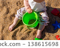 Toddler sitting on sandy beach holding green plastic bucket with sand toys around. Outdoor beach photography. Childhood and summer vacation concept. Design for poster, banner, postcard. Top view shot 138615698