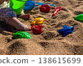 Toddler legs with plastic bucket and colorful sand molds on beach sand surface. Outdoor beach macro photography. Childhood play and summer vacation concept. Close up shot 138615699