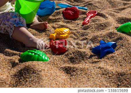 Toddler legs with plastic bucket and colorful sand molds on beach sand surface. Outdoor beach macro photography. Childhood play and summer vacation concept. Close up shot 138615699