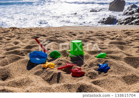 Plastic bucket with beach tools and molds placed on sand with sea and rocks in background. Outdoor beach photography. Summer vacation and seaside recreation concept. Wide angle shot 138615704
