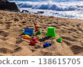 Plastic bucket with beach tools and molds placed on sand with sea and rocks in background. Outdoor beach photography. Summer vacation and seaside recreation concept. Wide angle shot 138615705